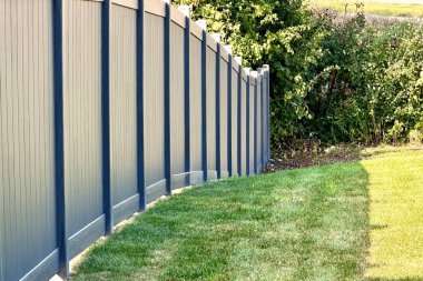 Güneşli bir günde yeşil çimenler ve ağaçlarla çevrili bir arazi sınırını gösteren modern bahçe çitleri ev sahipliği, sınırlar ve yerleşim alanlarını temsil ediyor..