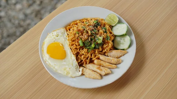 fried noodles with sunny side up eggs and pieces of chicken and cucumber on a white plate