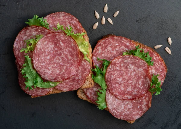 sliced salami and lettuce on black background, top view. open sandwiches with sliced salami sausage on rye bread