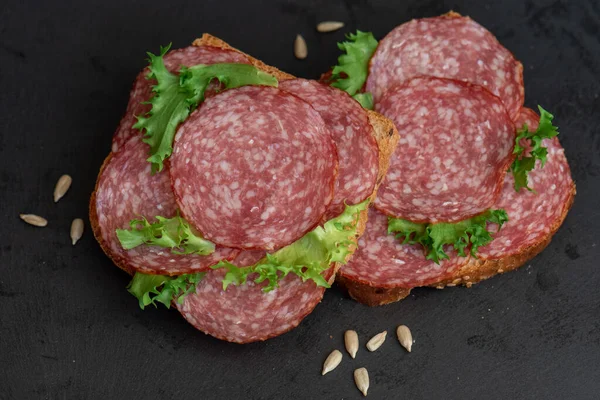 sliced salami and lettuce on black background, top view. open sandwiches with sliced salami sausage on rye bread
