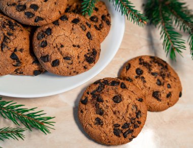 Tabakta çikolatalı kurabiyeler ve masada ladin dalları. Tabakta siyah çikolatalı kurabiyeler, Noel kurabiyeleri çikolatalı tarçınlı tatlı bayramı, yeni yıl yemeği ve Noel yemeği.
