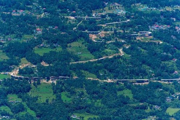 Yoğun yeşillik ve küçük binalarla çevrili yeşil bir tepe boyunca kıvrılan ve sakin ve resimli kırsal bir ortam yakalayan yılansı bir yolun manzarası..