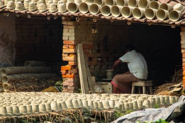 Bir zanaatkar, geleneksel bir açık hava atölyesinde el yapımı çömlek üzerinde çalışır. Kurutulmuş kil kaplarıyla çevrili kültürel zanaatkarlık sergiler..