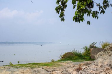Kıyıda yeşillik, kayalık bir sahil ve ufka uzanan sakin suların yer aldığı Ganga nehri manzarası..