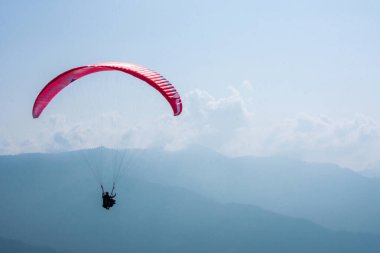 Renkli bir yamaç paraşütü gökyüzünde huzur içinde süzülüyor, nefes kesen bir dağ ve bulutlarla dolu bir arka plan, özgürlük ve macera hissi veriyor..