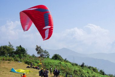 Canlı kırmızı bir para-planör fırlatmaya hazırlanıyor, yemyeşil bir yamaçla çevrili ve insanlar yardım ediyor. Dağ manzarası ve bulutlu gökyüzü manzaralı bir açık hava macerası ortamı yaratır..