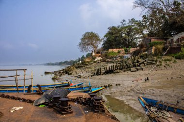 Çamurlu teknelerle dolu sakin bir nehir kıyısı, yıpranmış bir rıhtım, sudaki huzur ve tarihi tasvir ediyor..