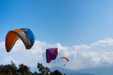 Parlak, açık mavi gökyüzünün altında yemyeşil ağaçların ve dağların üzerinde süzülen üç renkli yamaç paraşütçüsünün olduğu dinamik bir sahne. Hava sporlarının ve çarpıcı manzaraların güzelliği.