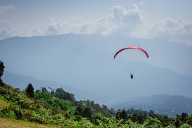 Renkli bir yamaç paraşütü gökyüzünde huzur içinde süzülüyor, nefes kesen bir dağ ve bulutlarla dolu bir arka plan, özgürlük ve macera hissi veriyor..