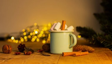 Beyaz bardaklı Noel afişi sıcak çikolatalı ve tarçın çubuklu marşmelovlu ve güzel çelenkli köknar ağacı dallı..