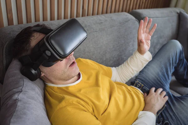 People and technology metaverse concept. a man watching a video in virtual reality glasses lying on the couch at home. 360 degree video viewing