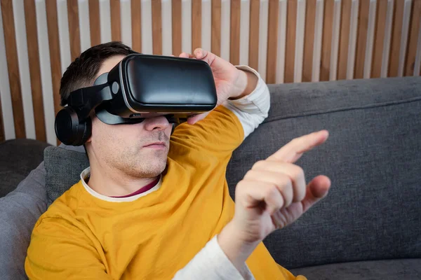 Smiling mature Adult male wearing VR glasses presses by his finger on virtual reality interface at home, game, technology, metaverse, viewing 360 video
