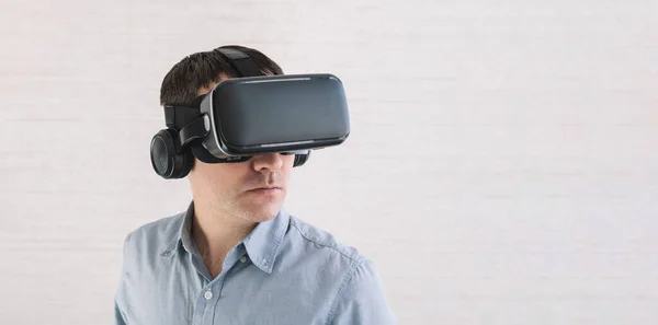 stock image Astonished caucasian male in blue t-shirt, using oculus rift headset, experiencing virtual reality while playing video game, looking joyful, white wall. 360 degree video viewing