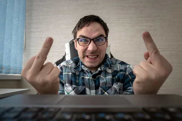 Stock image a man writes comments while sitting at a laptop and shows his middle finger at a computer screen monitor. insulting and bullying in comments and chats on the Internet. Fuck you sign.