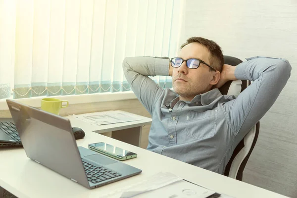Happy relaxed Caucasian young male rest in chair distracted from computer work, relieve negative emotions. Calm millennial man in glasses sit relax at home office workplace take nap or daydream.