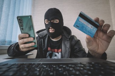 Soyguncu maskeli erkek hacker, sahte planda telefon, kredi kartı ve dizüstü bilgisayar kullanıyor. Siber hırsız kişisel verileri ve kredi kartı bilgilerini çaldı. Hacker kullanıcının parasını çalmak için kötü amaçlı yazılım kullanır.