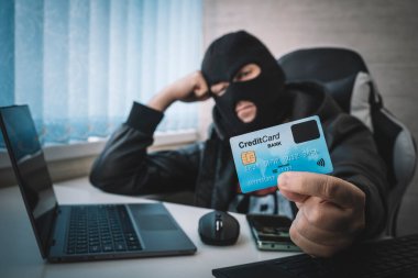 İnternet Hırsızlığı - kar maskesi takan ve elinde kredi kartı olan bir adam dizüstü bilgisayarın arkasında oturuyor, beyaz bir geçmişi var.