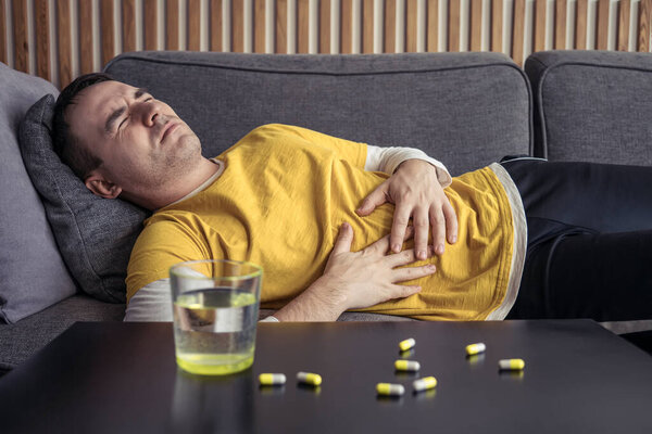 A man in home clothes and holds his hands on his stomach lying on the couch. the table, on which there are tablets, pills and a glass of water. Severe abdominal pain, malaise, cold.