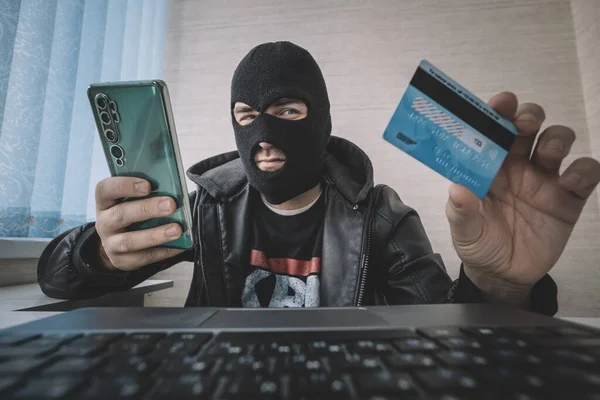 Male hacker in a robber mask uses phone, credit card and laptop in some fraudulent scheme. Cyber thief stole the personal data and credit card information. Hacker uses malware to steal user\'s money.