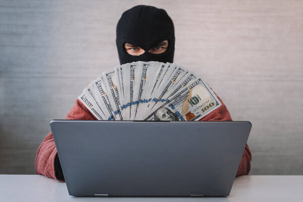a male thief holds dollars in his hand. using a laptop computer for password hacking activities. Cyber crime concepts.