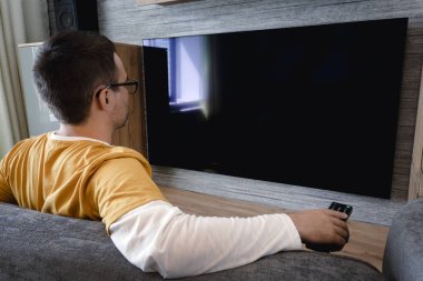 Adam televizyon izliyor, plazma düz ekran TV açıyor, beyaz duvardaki boş TV ekranına uzaktan kumanda doğrultuyor. Adam evde kanal değiştiriyor, arkadan bakıyor. Model