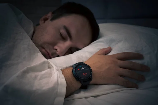 Fitness Activity Tracker With Heartbeat Rate On Man's Hand Over Bed. a man sleeps on a bed with a heart rate check in his sleep. Smart watches measure the pulse on the hand of a sleeping person.