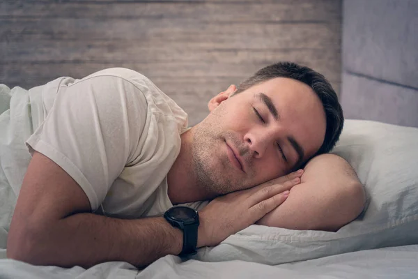 sleeping beautiful man. A satisfied expression on his face. A young guy is resting in bed with a smartwatch on his hands.