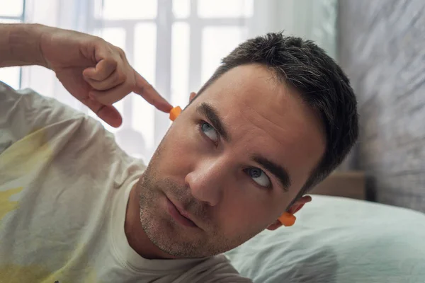 Attractive man in his 30s putting on earplugs before sleeping again because of a noisy morning and neighbors. plug in the ears for silence during sleep. noisy neighbors interfere with sleep
