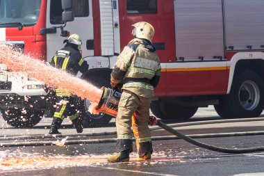 İtfaiyeci - İtfaiyeciler büyük bir yangını söndürüyorlar, koruyucu giysileriyle itfaiye aracının arka planında duruyorlar. İtfaiyeciler yangın söndürücü kullanıyor.