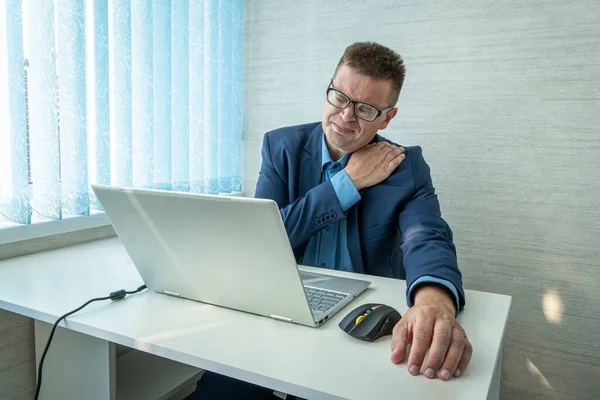 man with stiff shoulder in front laptop. health problems from sedentary work. a man in a suit is holding on to a sore neck