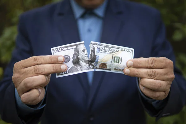 Close-up of human hands tearing hundred-dollar banknote. The concept of losing money and income. A businessman in a jacket tears up a paper bill of 100 US dollars