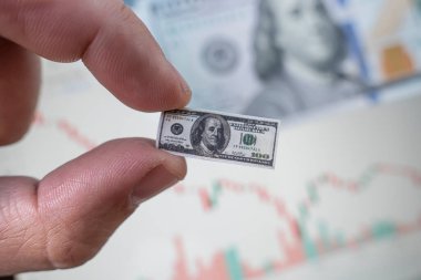 Borsada düşmekte olan bir tablonun arka planına karşı 100 dolarlık küçük bir banknot tutuyor. Borsa ticaretinden düşük gelir. İflas Doları Enflasyonu.