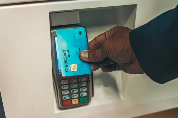 Contactless Payment System. Afro guy in suit holding credit card close to electronic payment machine, paying for delivery. Man paying with NFC technology on credit card, restaurant, shop.