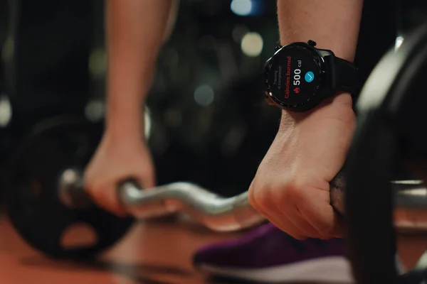 close-up of a smart watch on a woman\'s hand lifting a heavy barbell in the gym on a dark background. Burning calories. Technology and sports concept