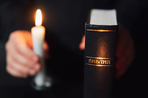 男人的手拿着圣经和两支燃烧的蜡烛在黑色背景下祈祷 这本书的题词翻译自俄文 — 图库照片