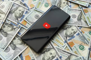 Akıllı telefon ekranı ve para arkaplanındaki mobil video sunucu uygulaması için Youtube logosu. Cep telefonu ve yüz dolarlık banknotlar, üst görüntü, yakın plan. 30 Mart 2023. Barnaul. Rusya.
