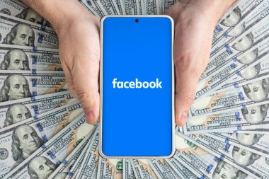Facebook uygulamalı akıllı telefon ekranı ve bir sürü yüz dolarlık banknot. İş ve sosyal ağ kavramı. 30 Mart 2023. Barnaul. Rusya.