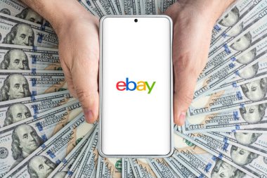 eBay web sitesinin logosu akıllı telefon ekranında erkeklerin elinde Amerikan dolarları üzerinde yatıyor. 30 Mart 2023. Barnaul. Rusya.