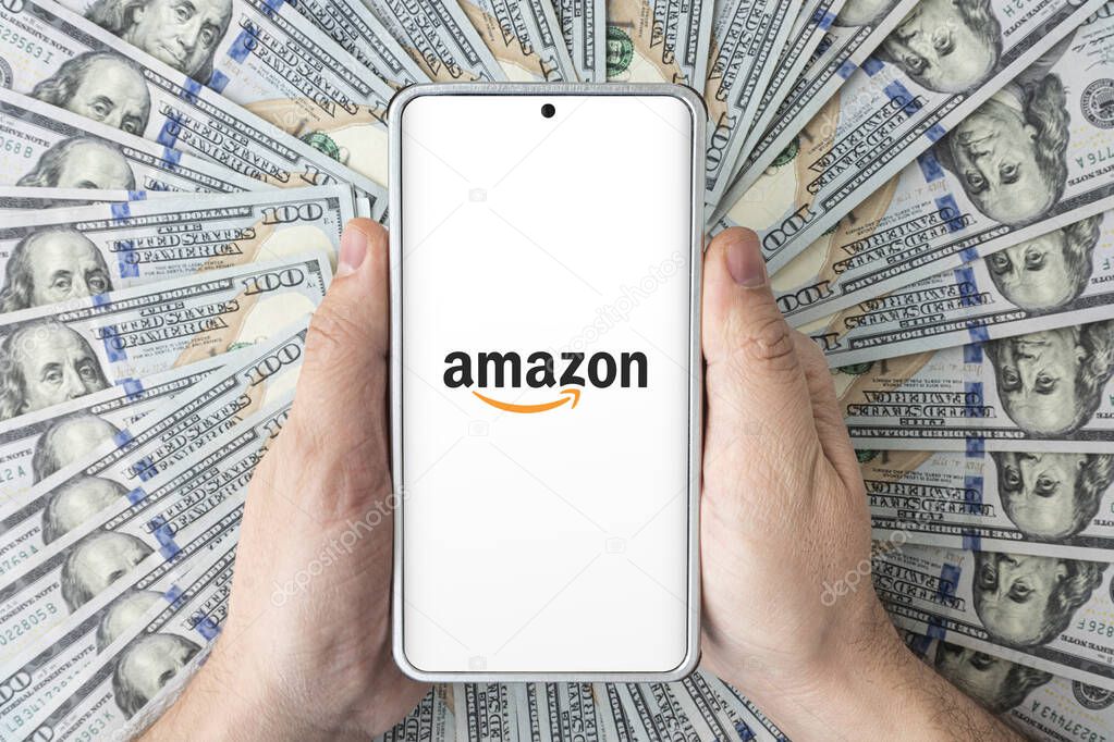 The logo of the amazon website on the smartphone screen in men's hands lying on American dollars. March 30, 2023. Barnaul. Russia.