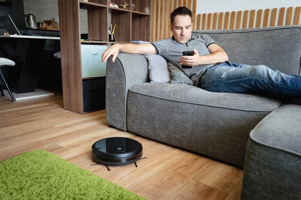 Robot vacuum cleaner cleaning the living room. man enjoy rest, sitting on sofa at home. Remote control of robot vacuum cleaner via smartphone. Smart technologies at home. Internet of Things concept