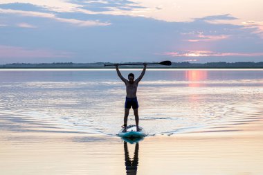 Günbatımında tek başına kürek çeken bir erkek silueti. Körfezde sörf tahtası. Gün batımında, genç bir adam, bir gölün üzerinde ayakta duran bir sörf tahtası ile kürek çekiyor..