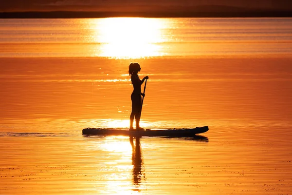 Girl on sup board with beautiful colored sunrise background. a young woman stands on a board with a paddle and swims on a beautiful lake