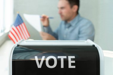 ABD 'de seçim. Bir erkek seçmen oy kullanma merkezinde oy veriyor. Oy sandığı ön planda Amerikan bayrağıyla birlikte.