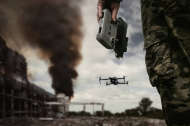 Uzaktan kumandalı İHA operatörü şehirde siyah dumanlı yıkılmış bir binanın arka planında duruyor. Modern savaşta insansız teknolojiler kavramı. bombardıman insansız hava aracı