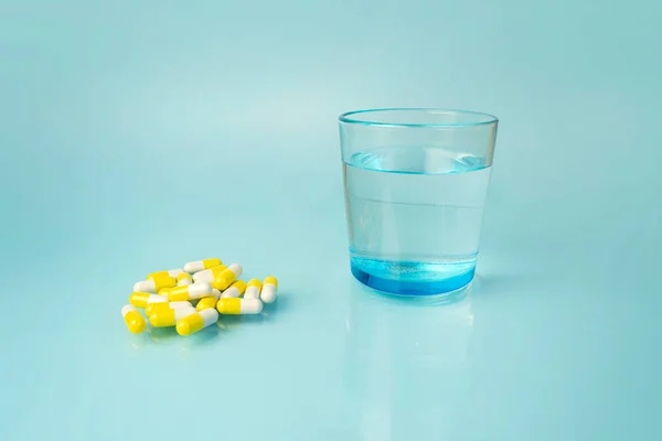 white and yellow medical pills, vitamins, in the distance there is a beautiful blue glass with clean water, on a harmonious bright blue background. Taking pills, vitamins. Selective focus.