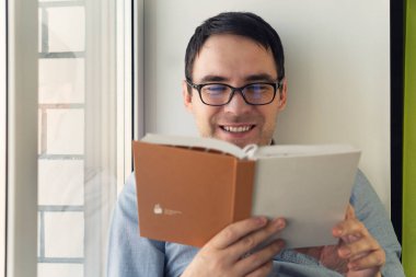 Evde kitap okuyan genç adam pencerenin yanında.