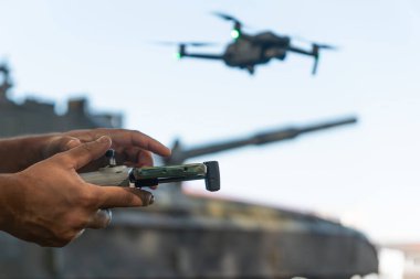 Elinde İHA kontrol paneli olan bir adam, arka planda tankın üzerinde uçan bir dron. Modern savaşta keşif dronlarının yardımı.