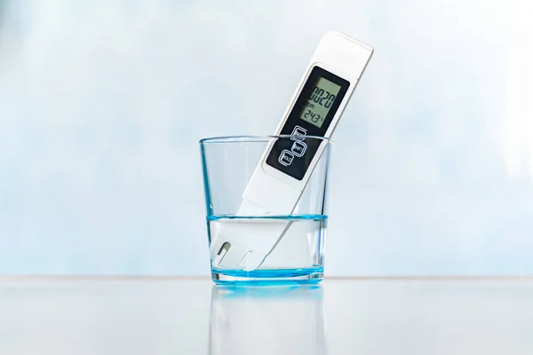 Water quality meter tester submerged in drinking glass, on blue background. Measuring harmful impurities, distilled water, purifying water for healthy eating.
