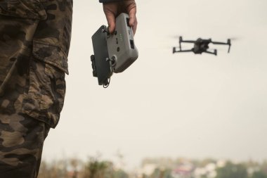 Elinde İHA kontrol paneli olan askeri üniformalı bir adam. Arka planda uçan bir dron.