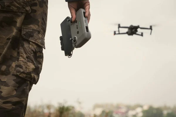 a man in a military uniform with a UAV control panel in his hands, a drone flying in the background in nature